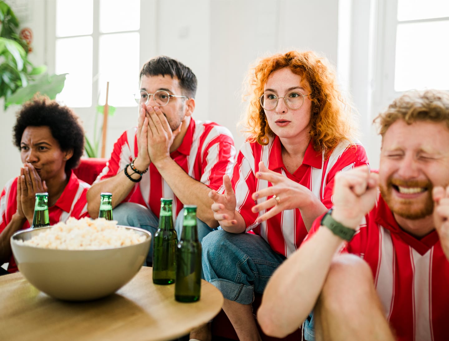 Amigos con camisetas de fútbol a rayas rojas y blancas, emocionados viendo un partido en la televisión. Friends wearing red and white striped soccer jerseys, excitedly watching a game on tv.