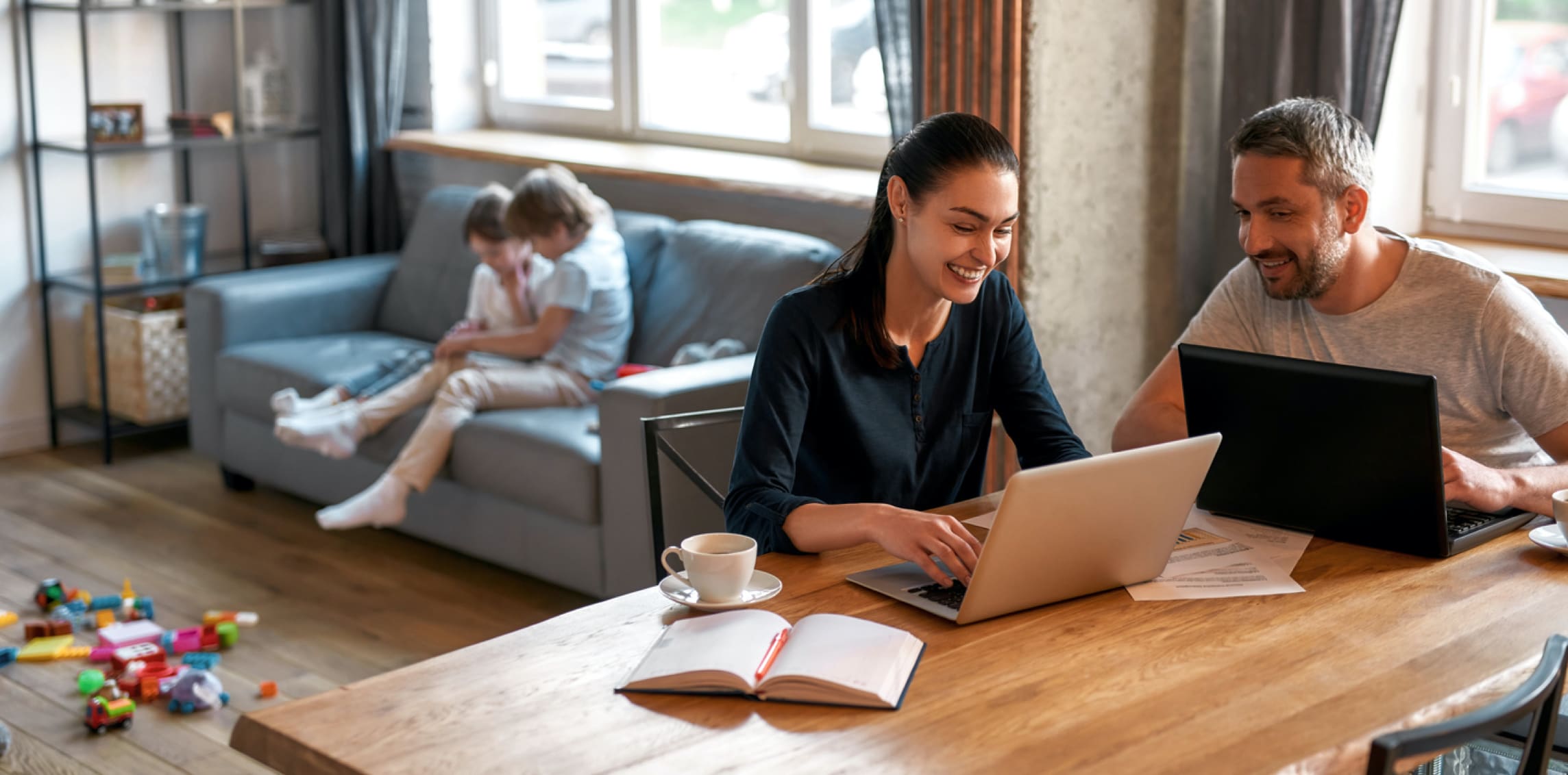 Husband and wife laugh together while both using laptops at a table and children play in the background.