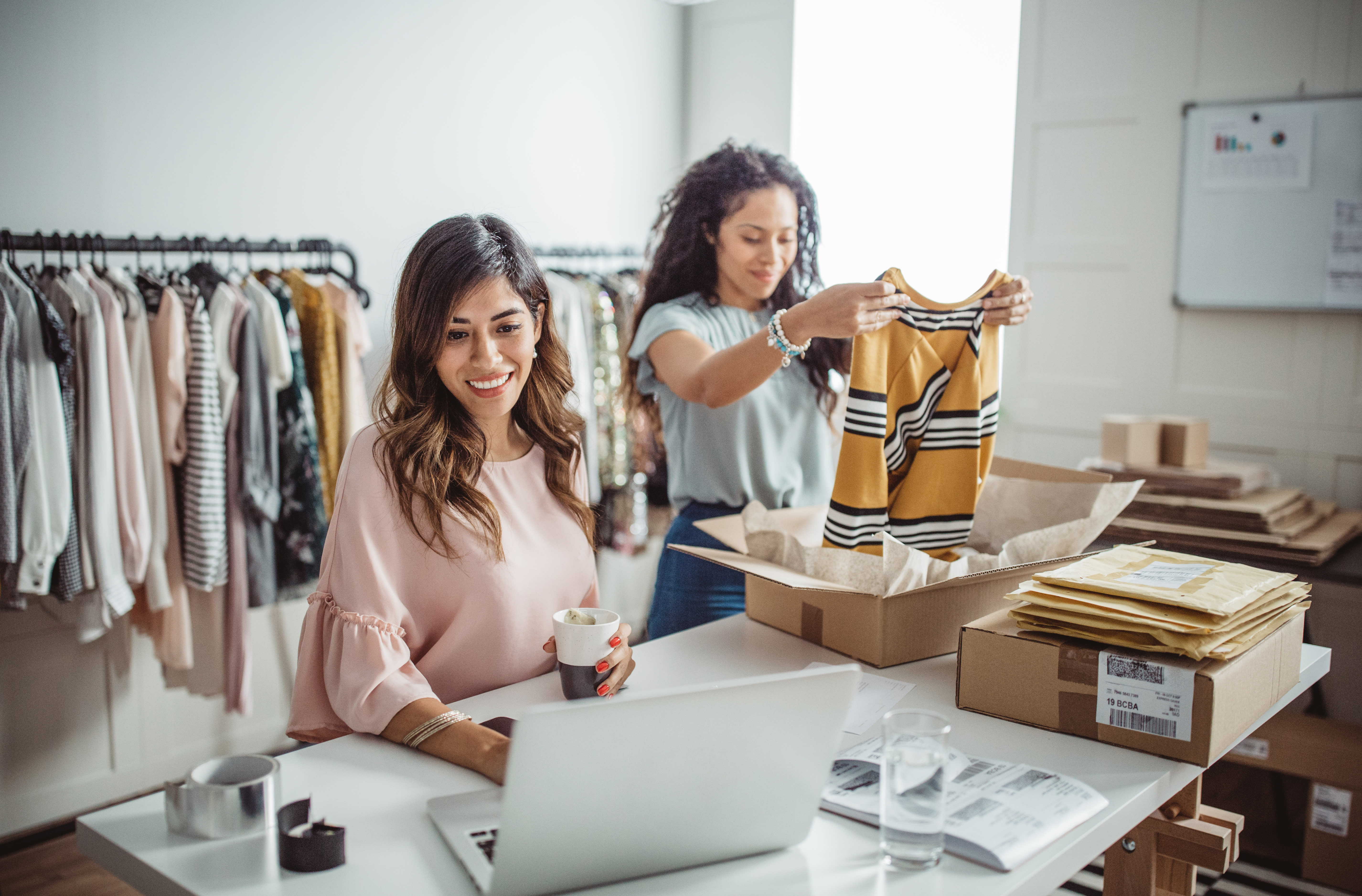 Two women packaging clothing. One of the woman is using a laptop.