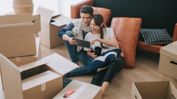 Couple sitting on the floor, surrounded by boxes. The couple is also looking at a tablet.