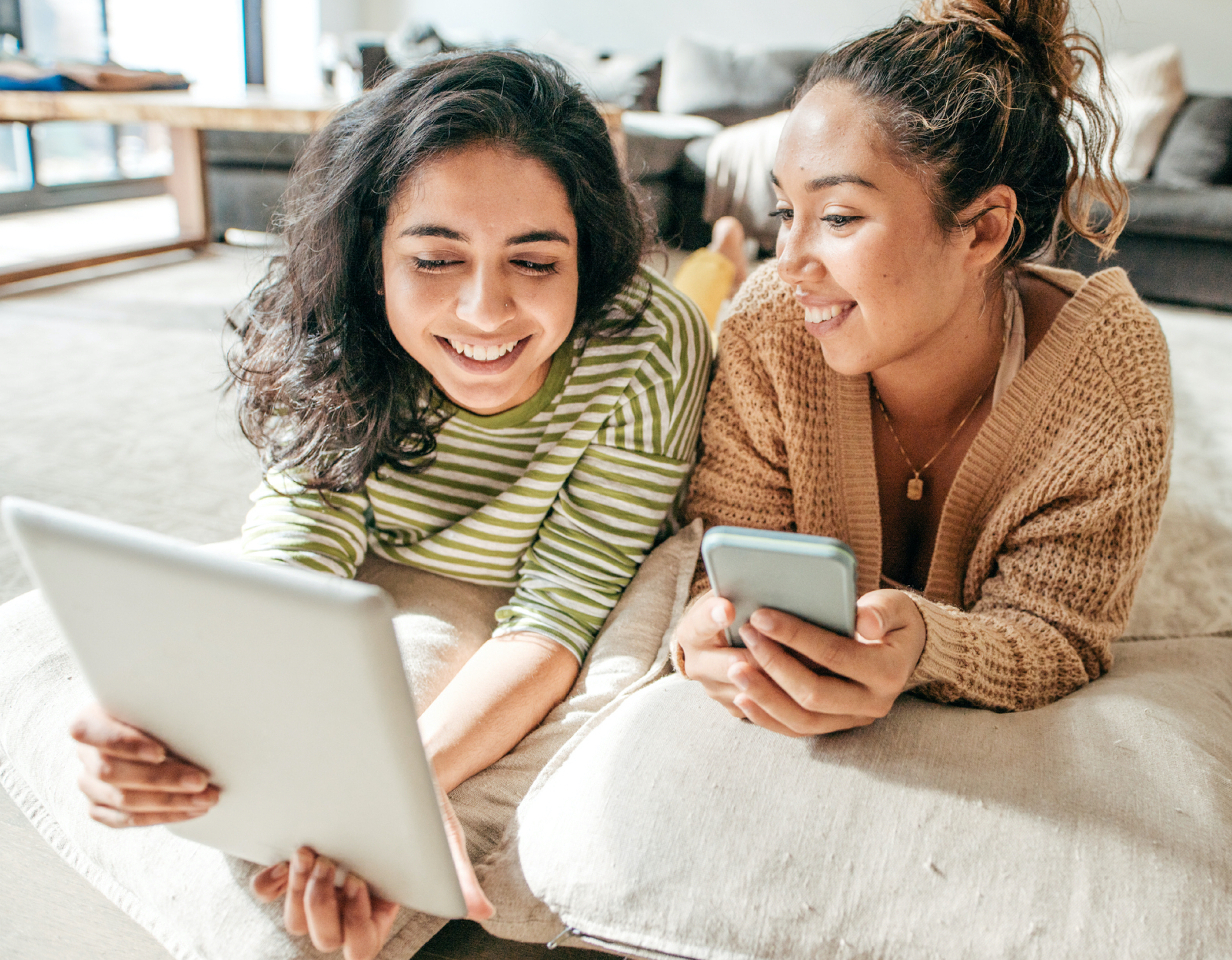 Two female friends smile while sitting next to each other with tablet and phone.