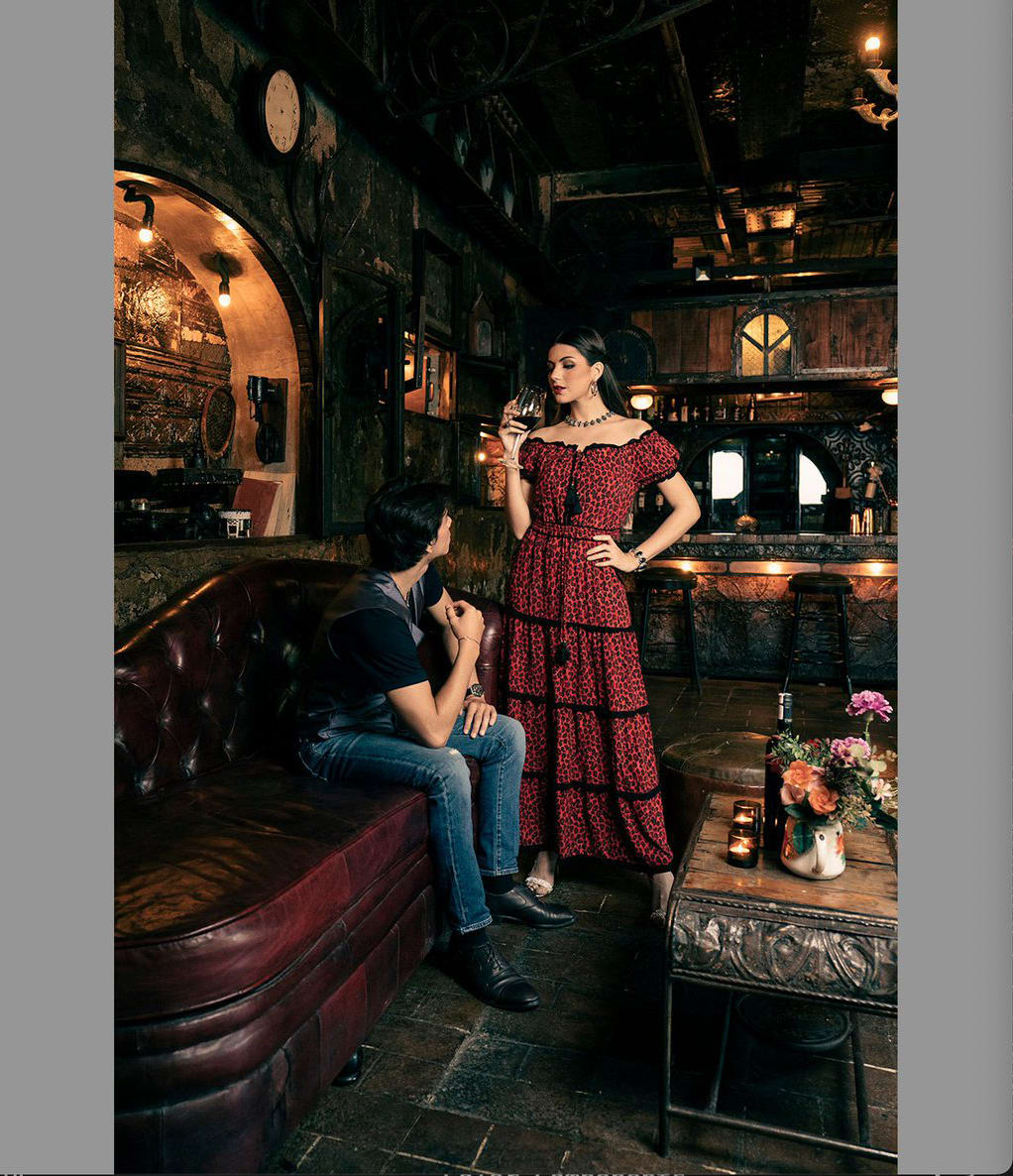 A woman in a tango red dress stands while holding a wine glass in her right hand. She looks down at a man sitting on a leather sofa. The photo is the same with the exception of a shift in the color of the dress.