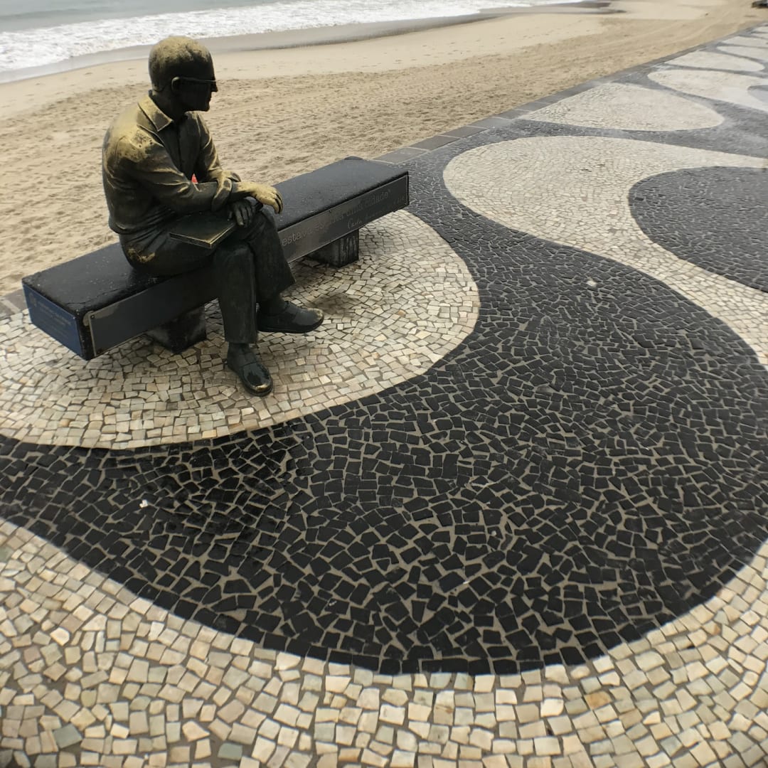 A statue of a man sitting on a bench is placed on a stone walkway near the ocean, with the ocean visible in the background.