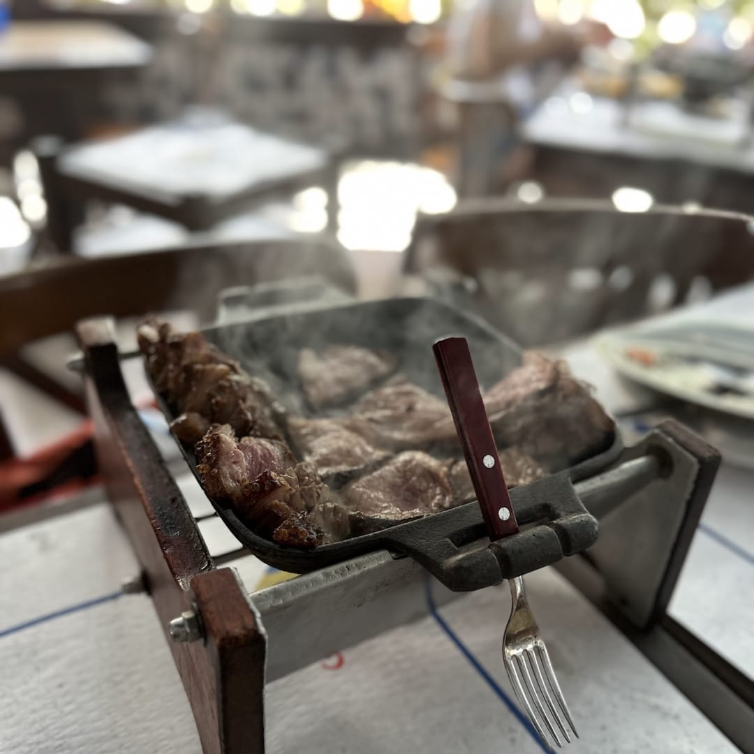 The image shows a dining table with a metal grill filled with various cuts of meat, including beef and pork, and a fork placed on the table.