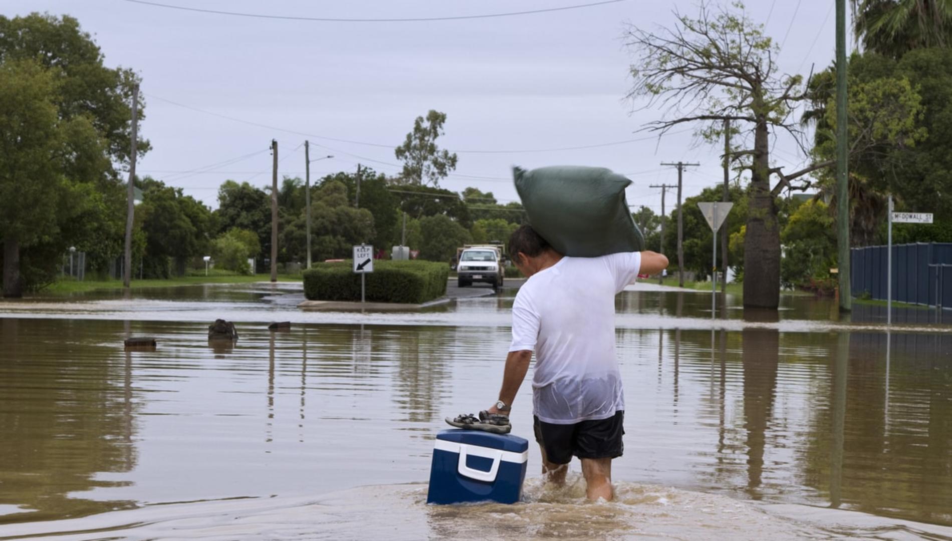 A man wading through floodwaters with a sack on his shoulder, pulling a floating Esky