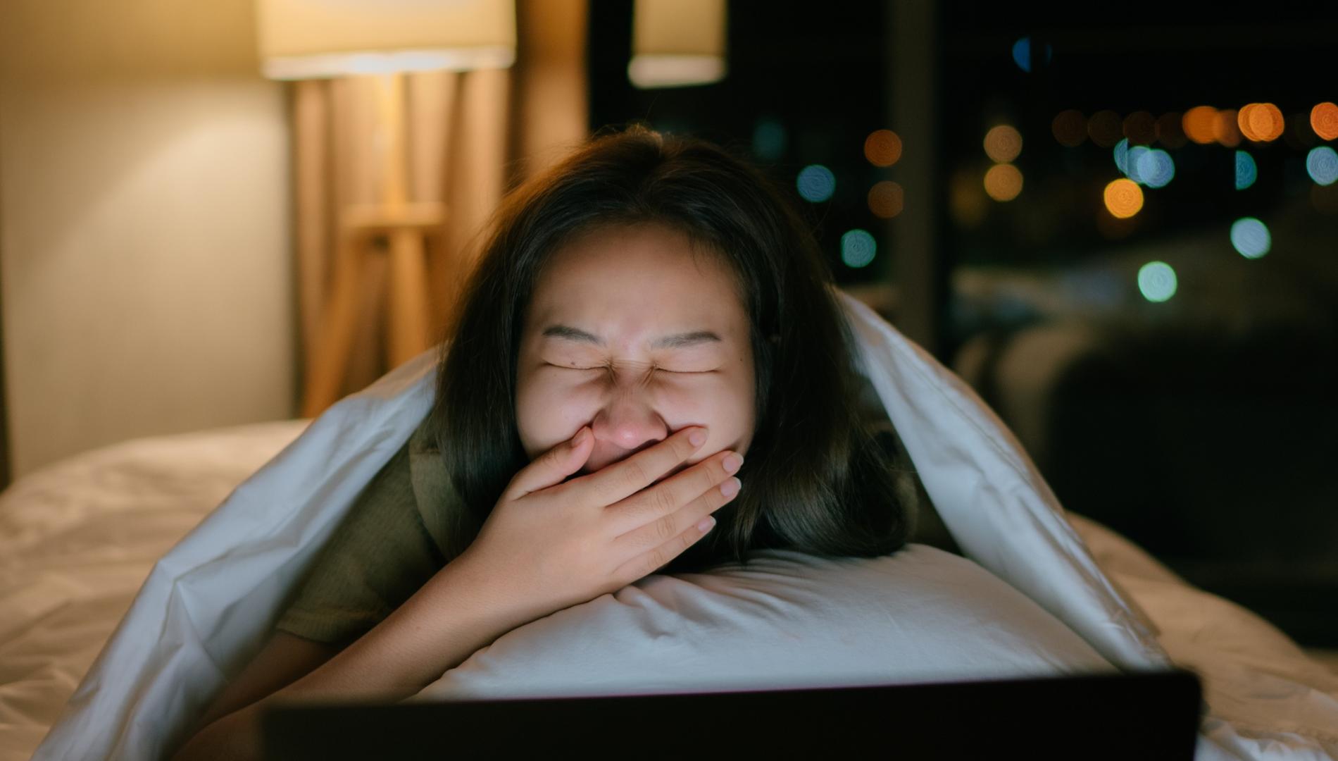 Woman lies on her bed with a computer, yawning