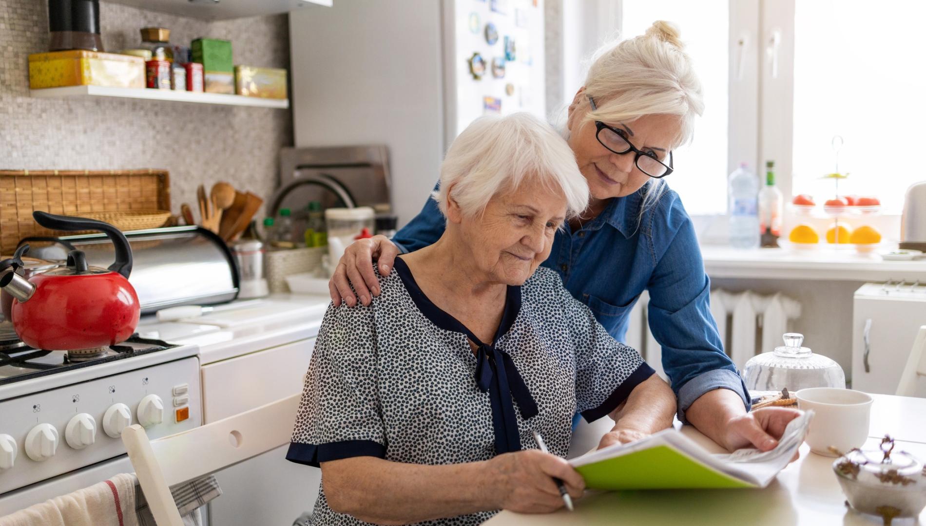 An elderly woman sits at a kitchen bench, with another women looking over her shoulder.