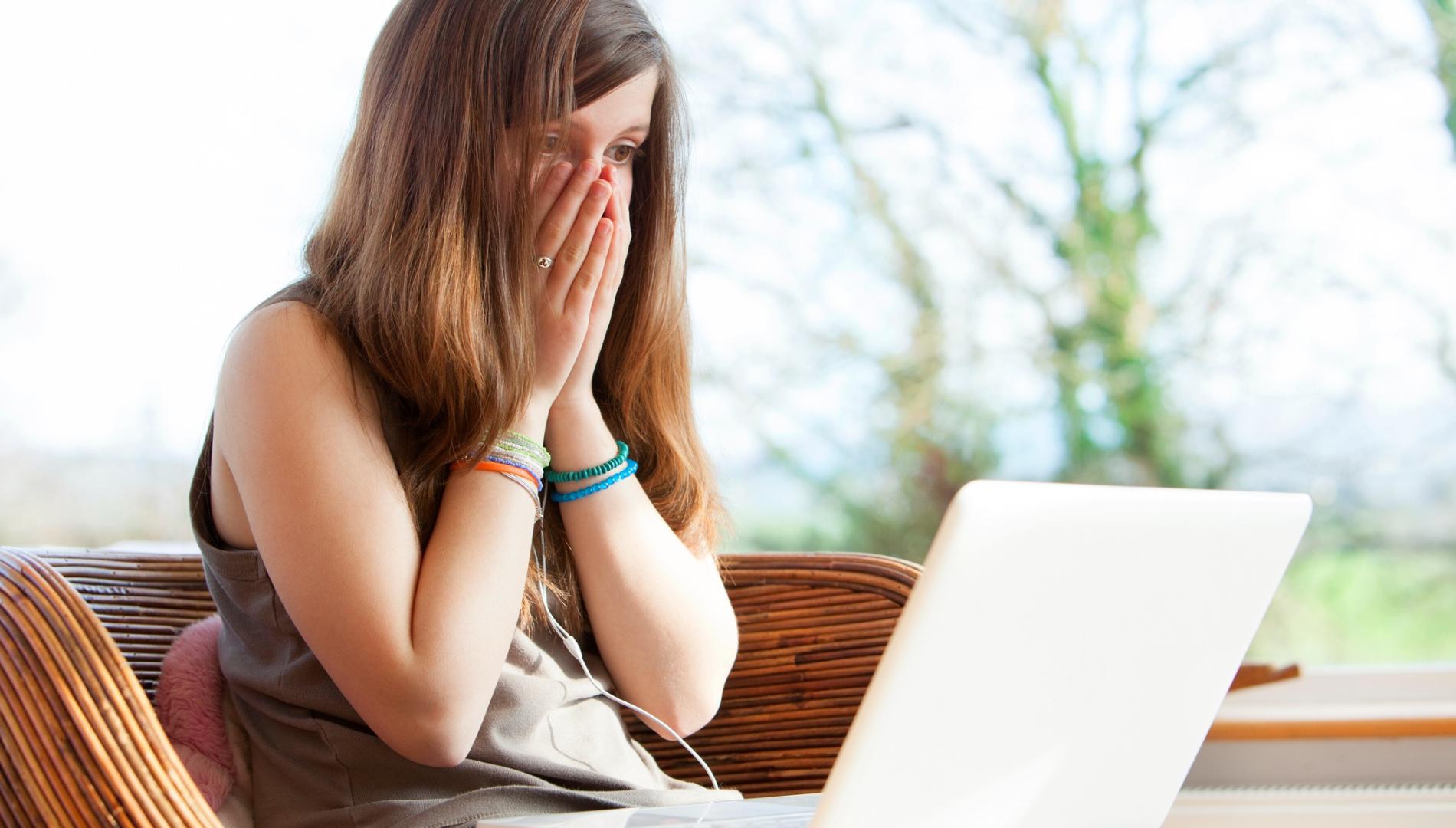 A young woman sits on a couch looking with shock at her laptop computer, her hands covering her face.