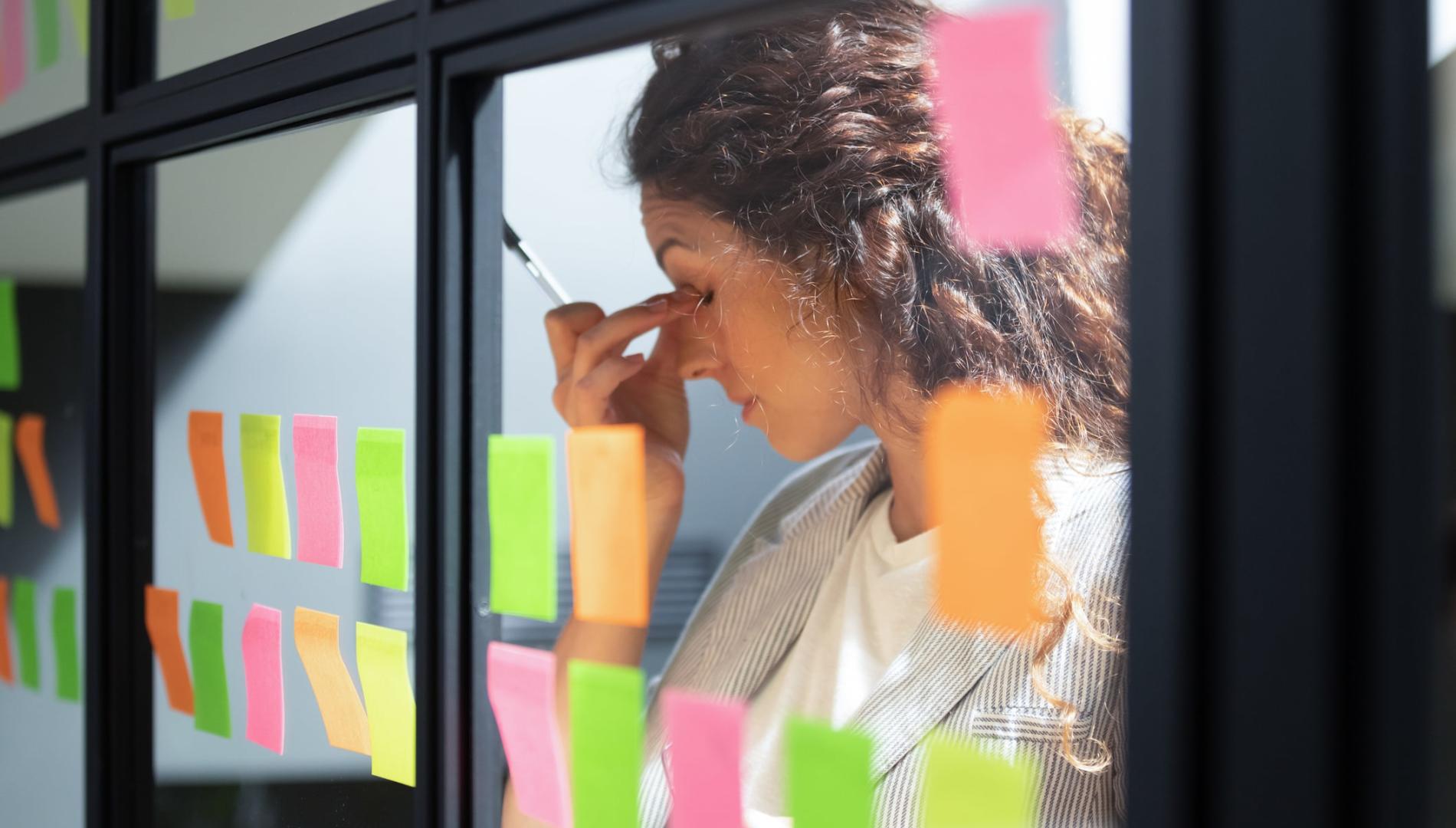 Tired woman touching bridge of her nose, working with sticky papers on windows