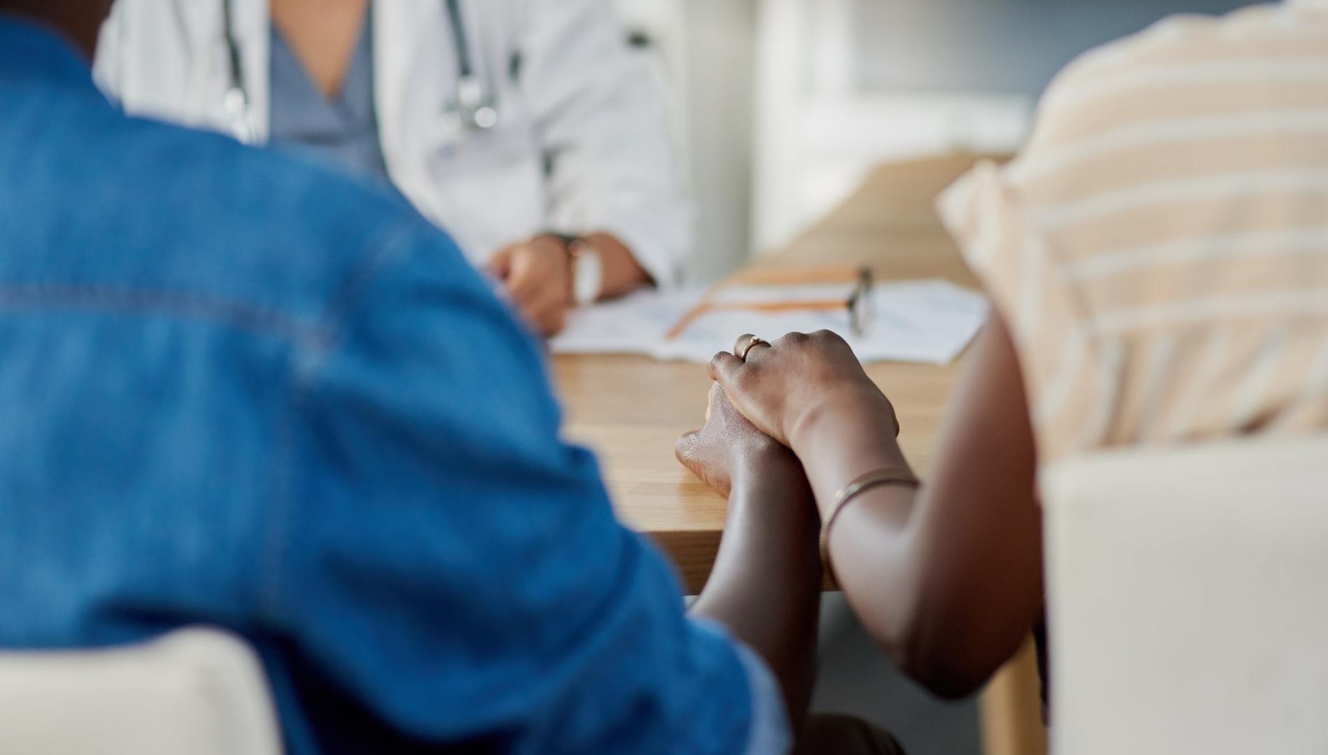 A man and woman seated in a doctor's office, holding hands, with the doctor behind a desk in the background