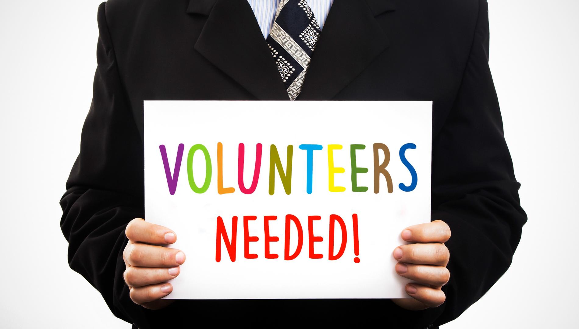 A man in a suit holds a sign with "Volunteers needed" written on it.