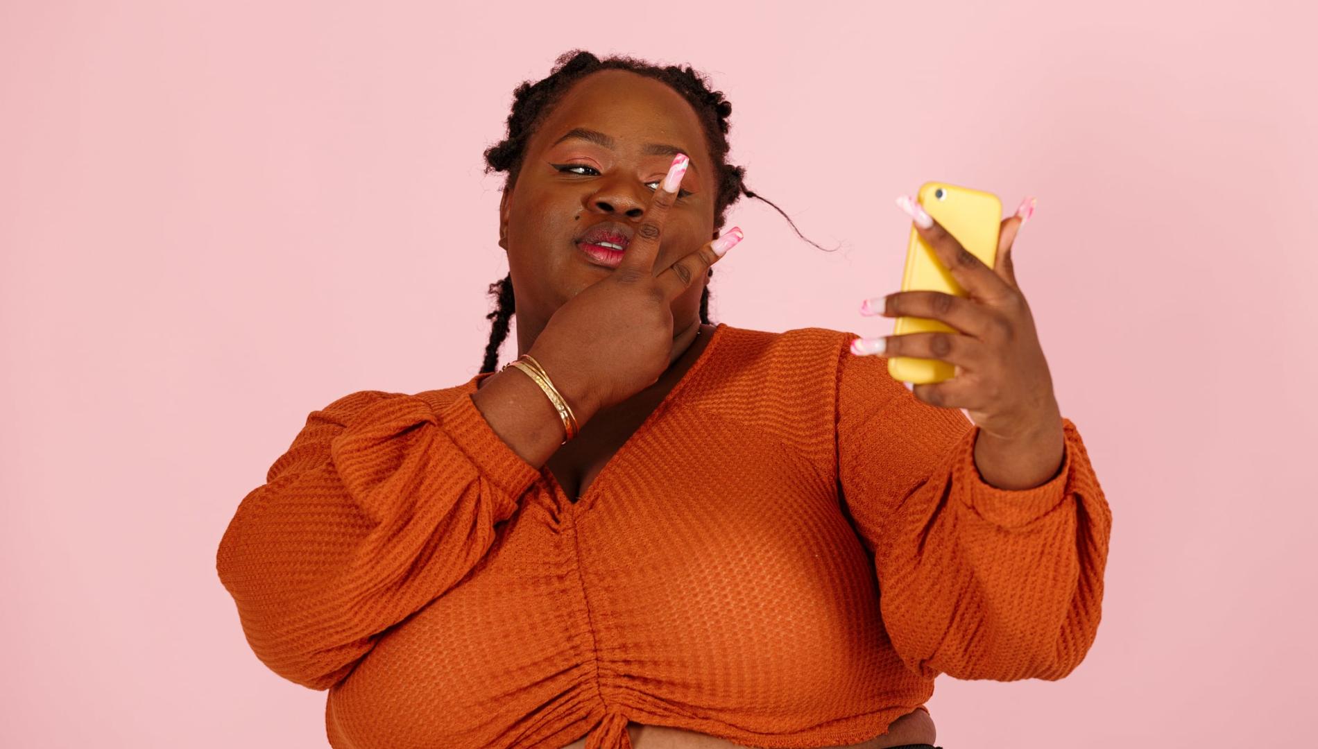 Funny young black woman taking a selfie with mobile phone on pink background
