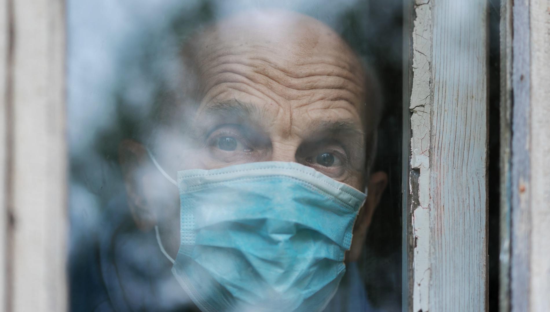 Sad-looking older man in medical face mask looking through a window