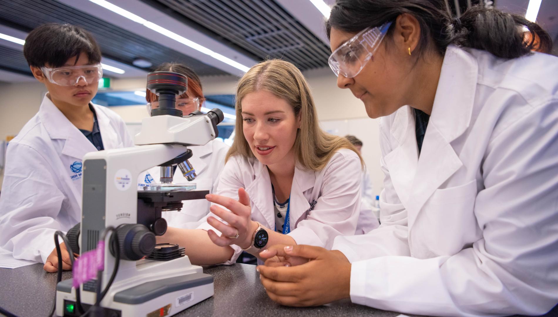 Laura Reid and students in lab coats and eye protection examining a microscope