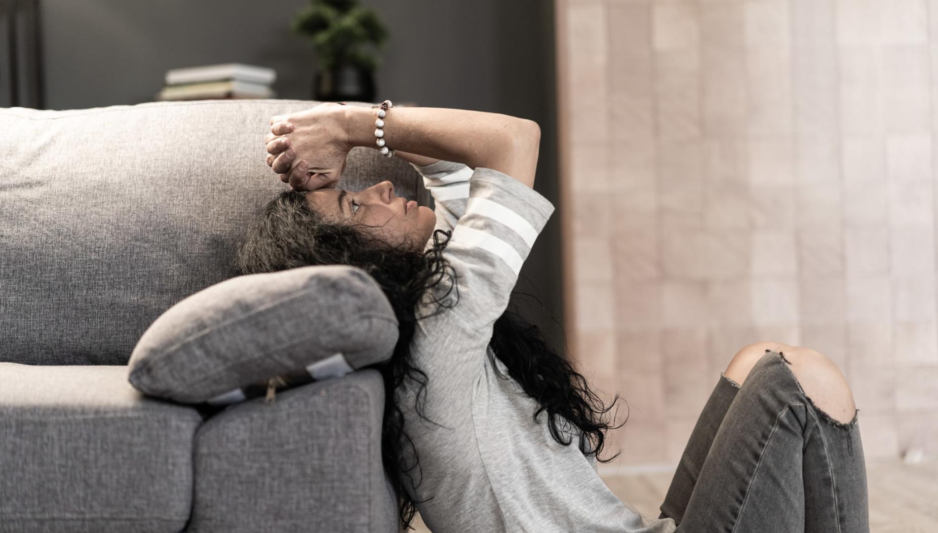 A despairing woman sitting on the floor and leaning against a sofa, with her hands on her head