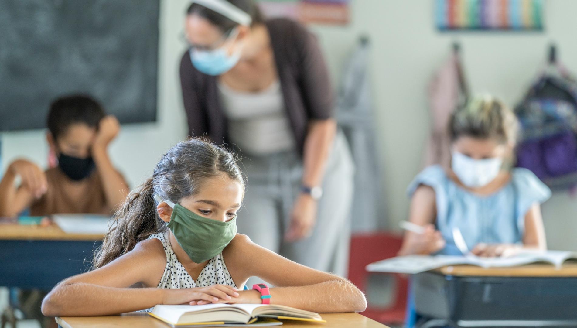 12 year old girl wearing a reusable, protective face mask in classroom while working on school work at her desk.