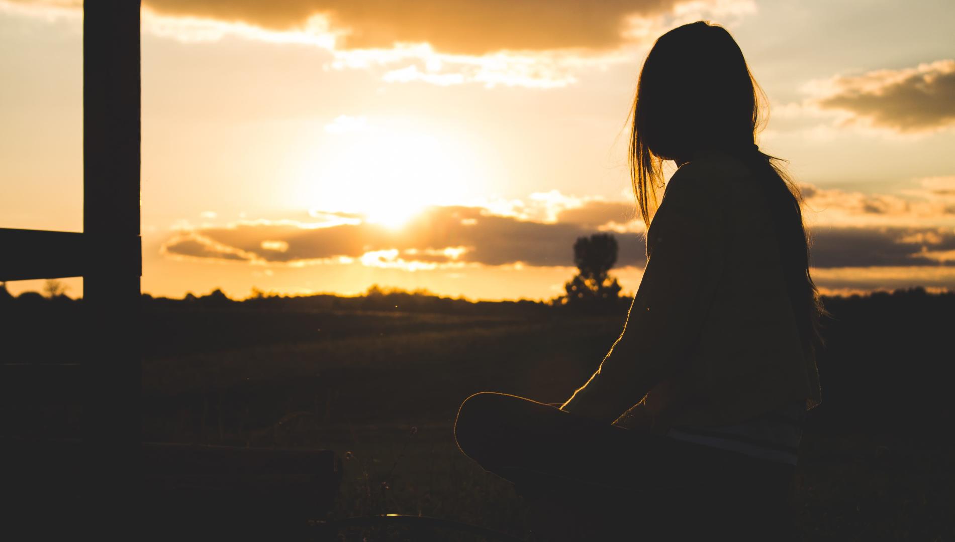 Silhouetted sitting woman gazing towards the sun behind clouds on the horizon