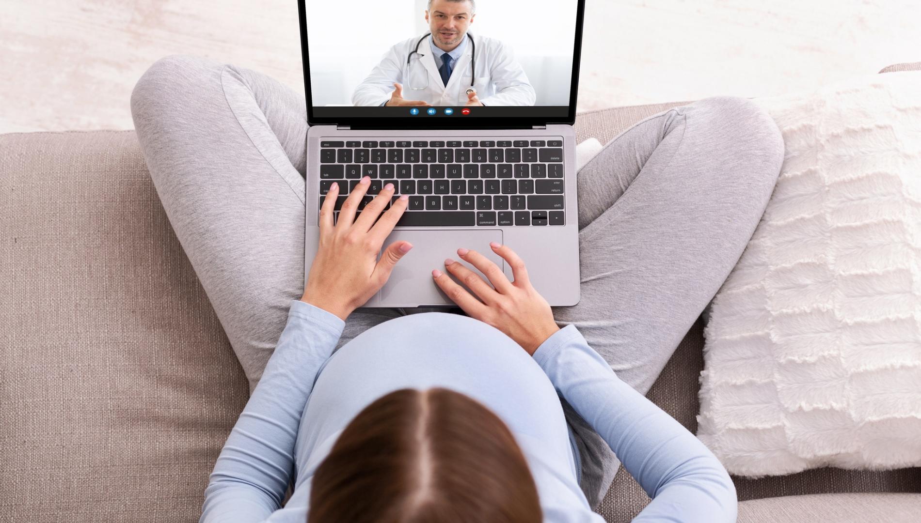 Pregnant women sitting on a couch on a telehealth video appointment with a doctor.