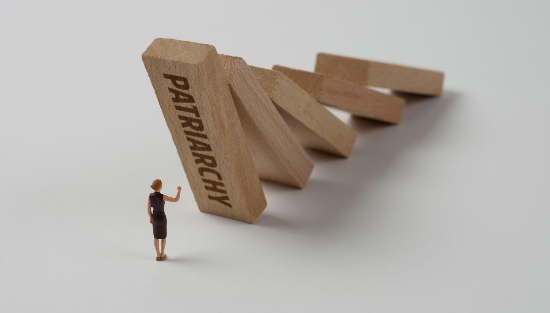 Female toy figure in front of falling wooden blocks with patriarchy written on one of them.