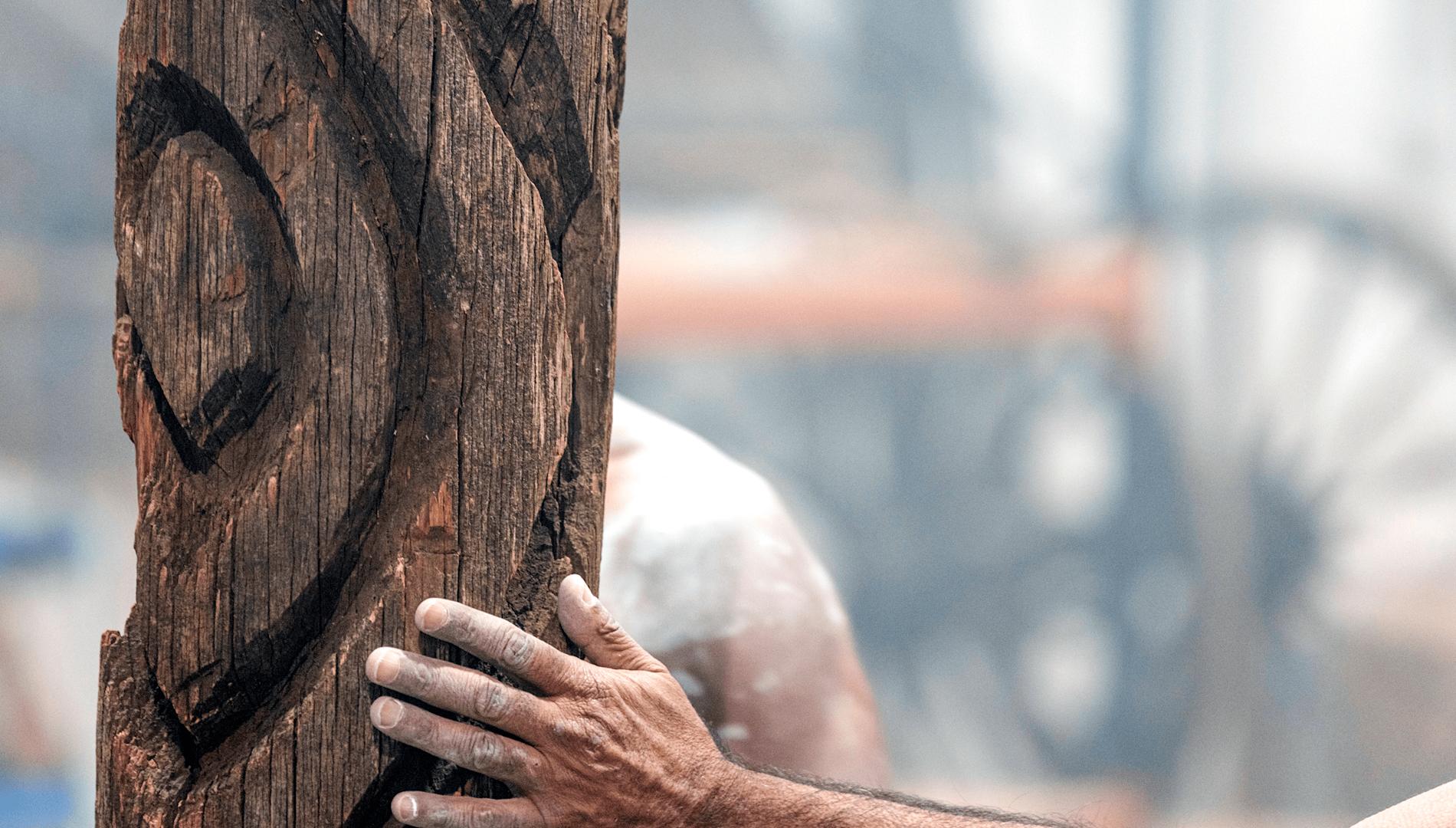 A carved sacred tree with a man's hand resting on it, smoke in the background