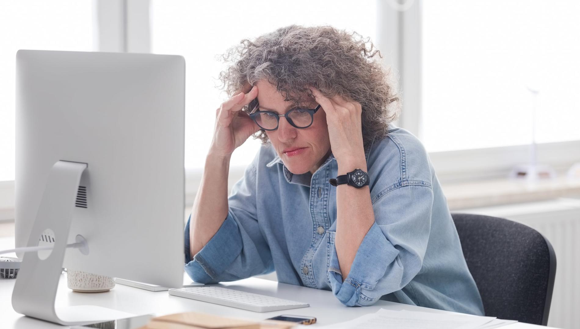 Older woman with hands on her head looking uncomfortable, sitting at a desk in front of a computer