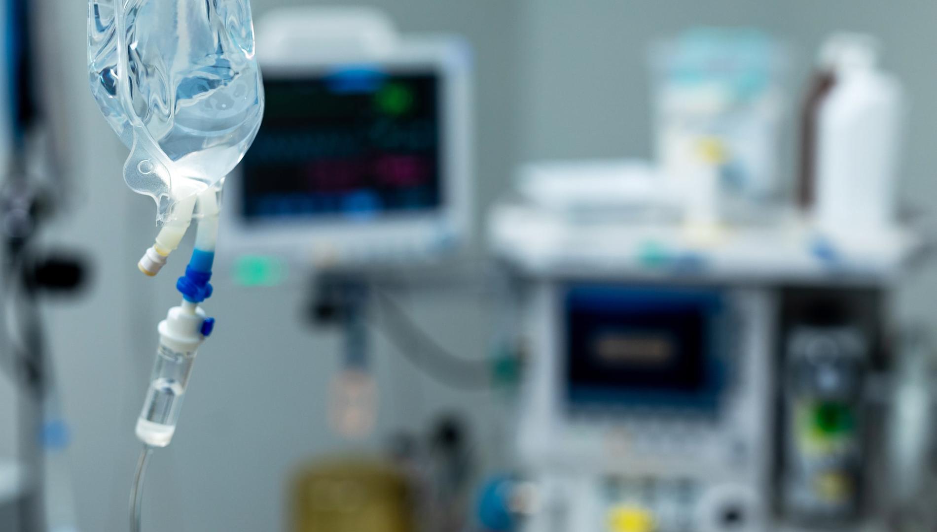 An IV drip bag with hospital equipment in the background