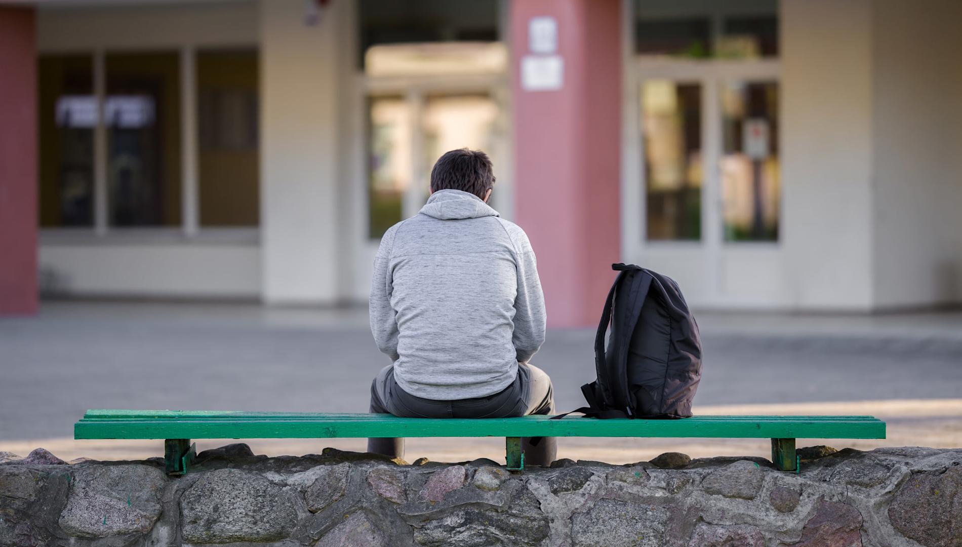 A young man with his back to the camera sitting on bench in a school yard