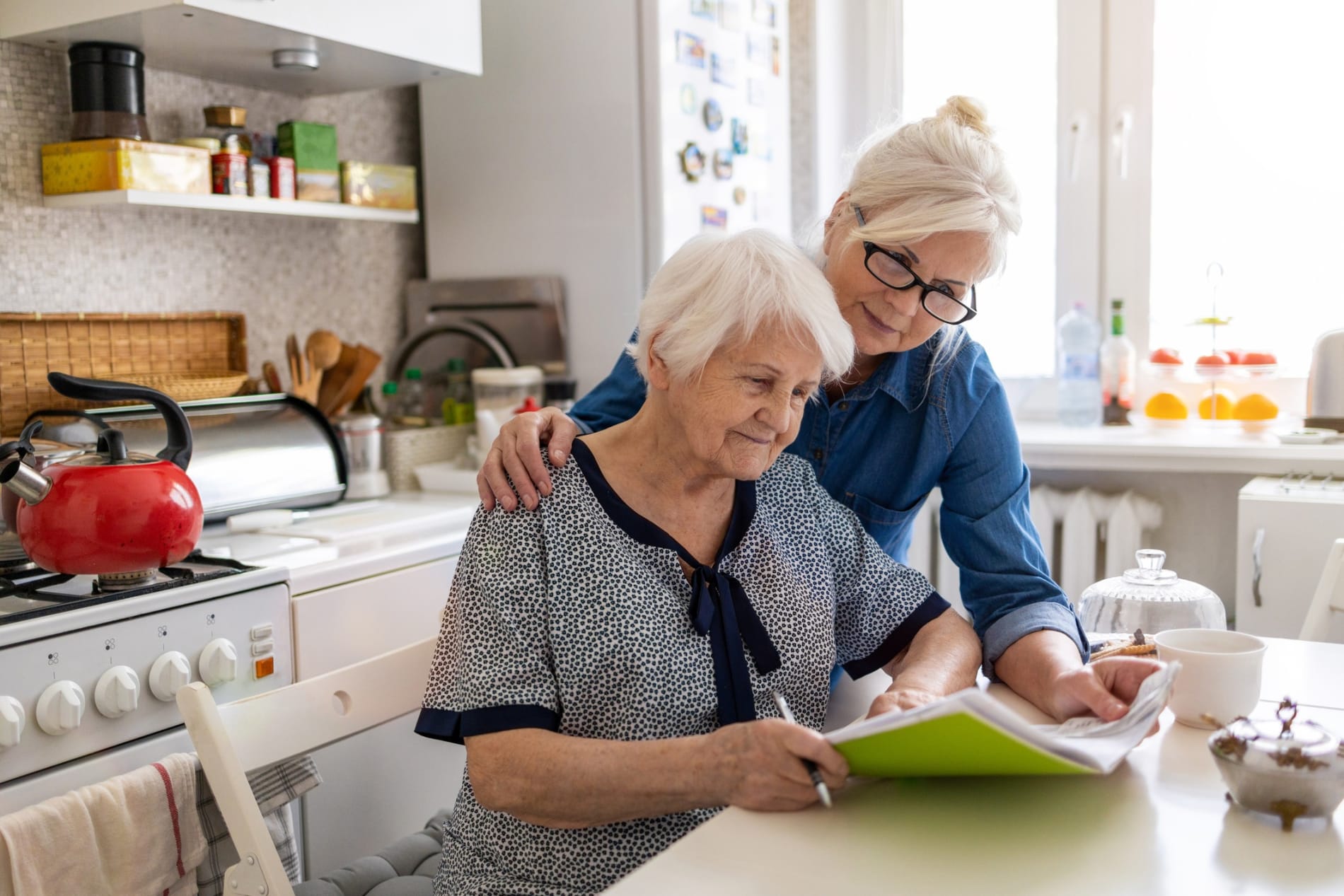 An elderly woman sits at a kitchen bench, with another women looking over her shoulder.