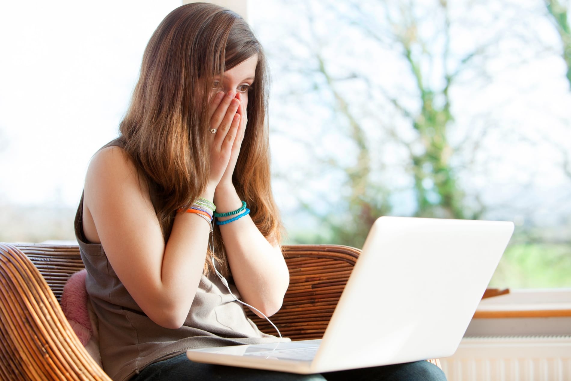 A young woman sits on a couch looking with shock at her laptop computer, her hands covering her face.