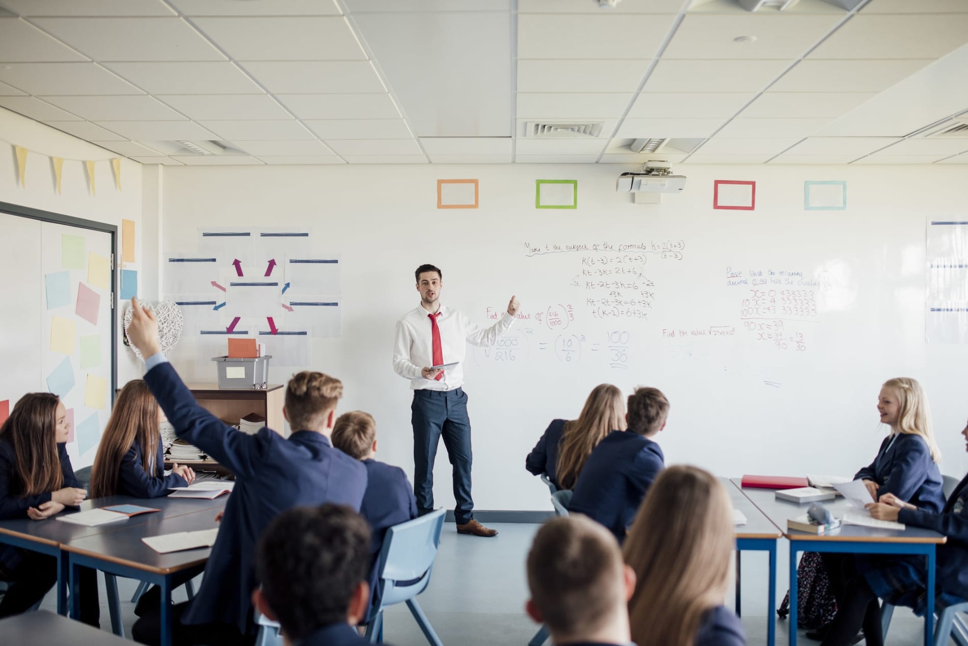 Young teacher at a whiteboard in front of students, one with hand raised