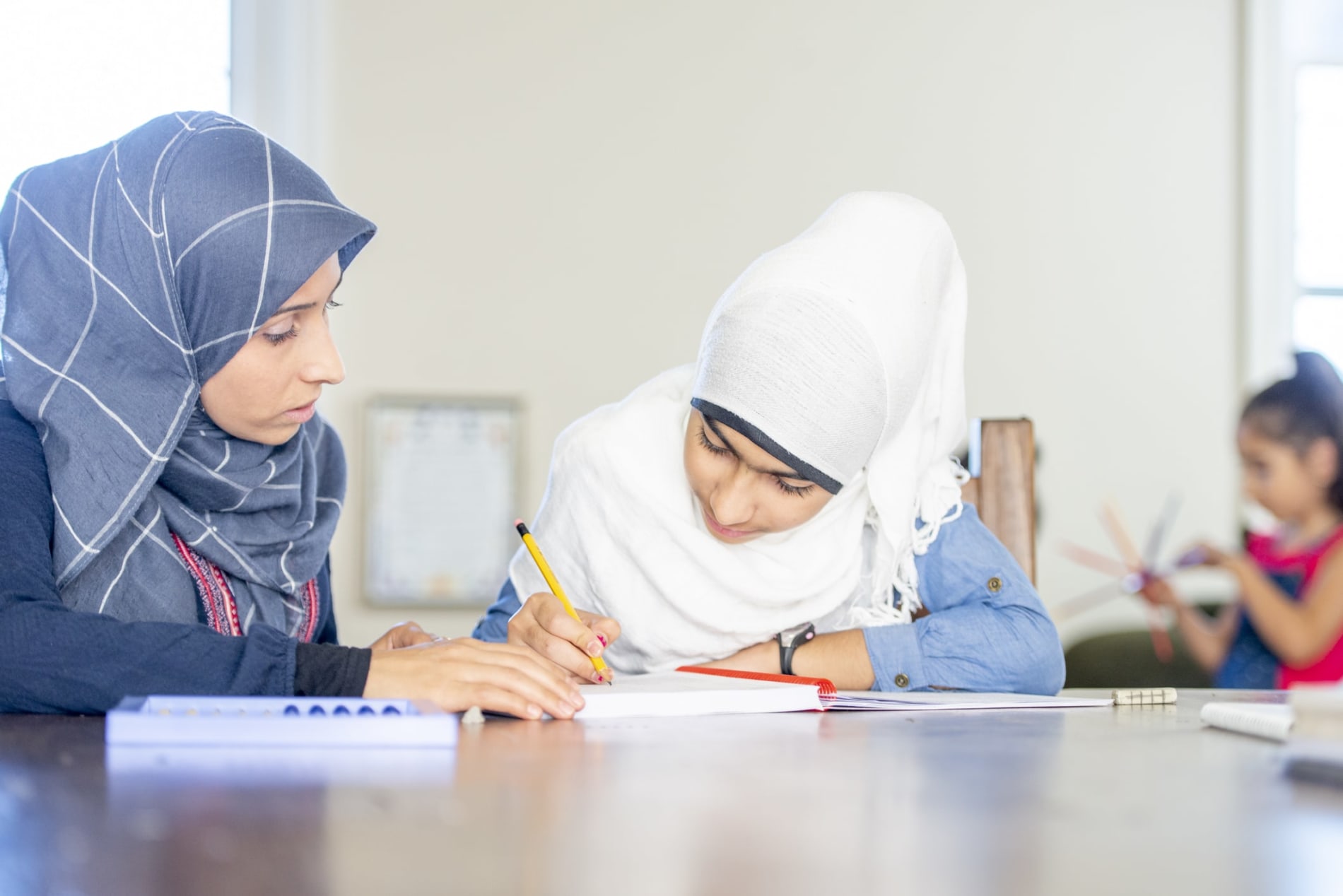 Teacher helping a girl with writing at a desk, both wearing hijabs 