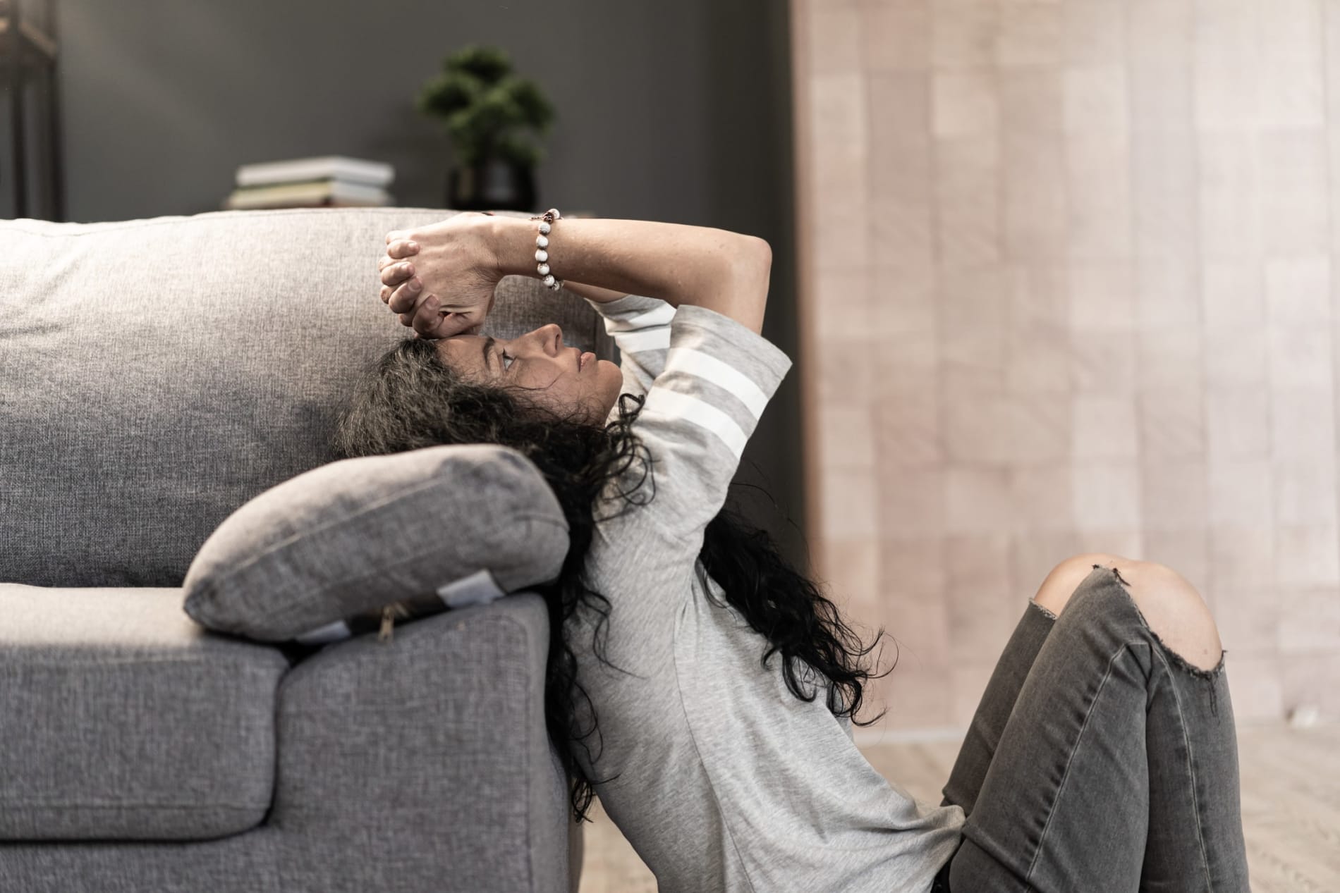 A despairing woman sitting on the floor and leaning against a sofa, with her hands on her head