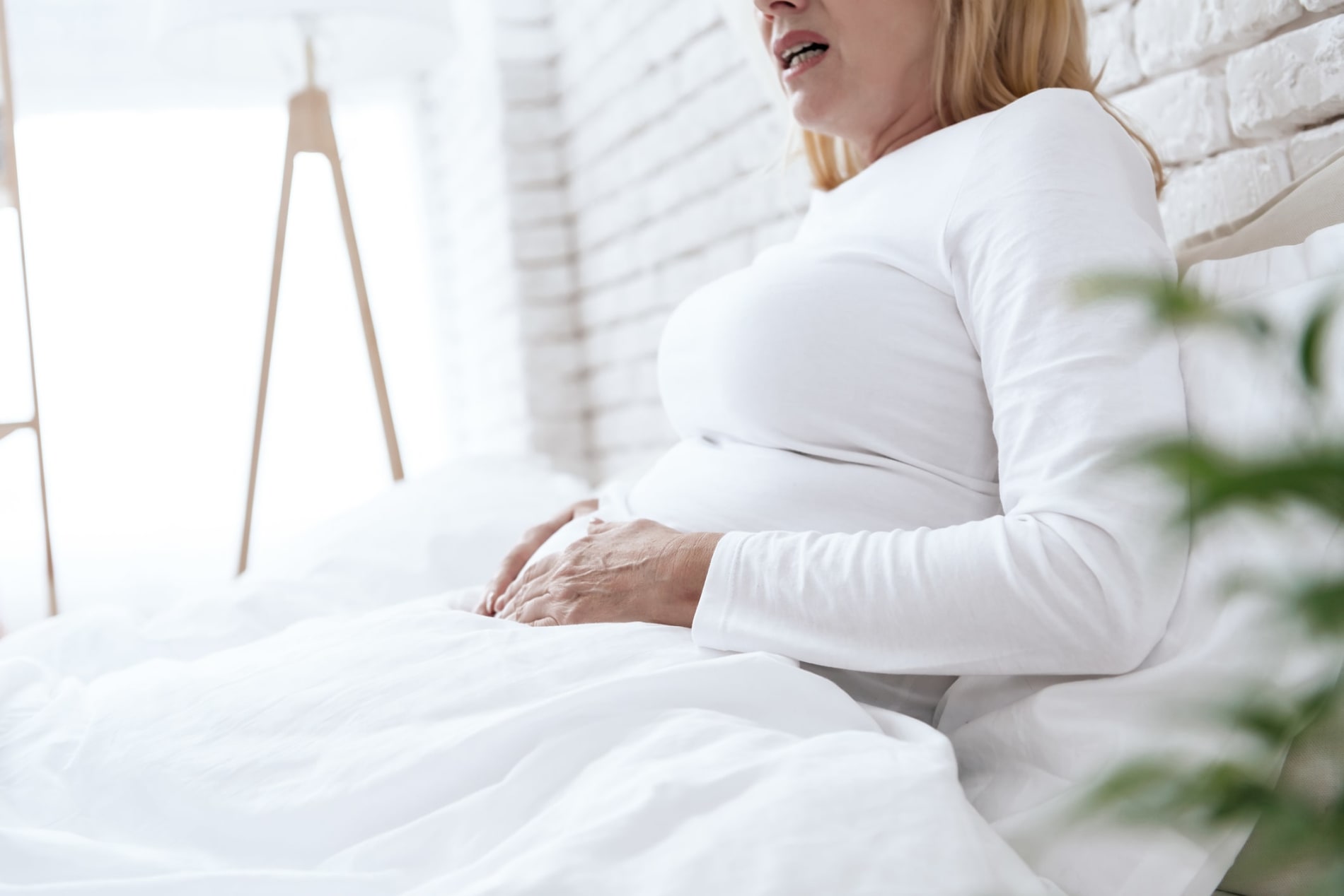 A woman in white clothing lying in bed clutching her stomach in discomfort