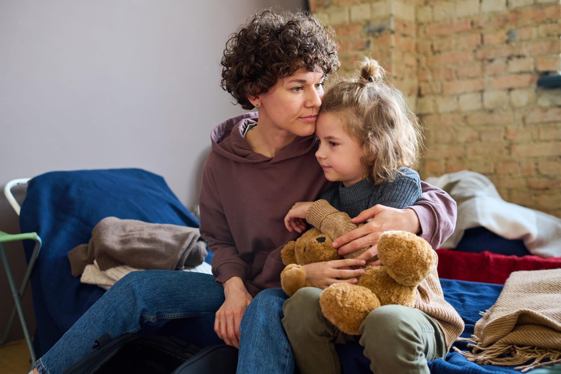 Young woman giving her young daughter a hug, who is holding a teddy bear.