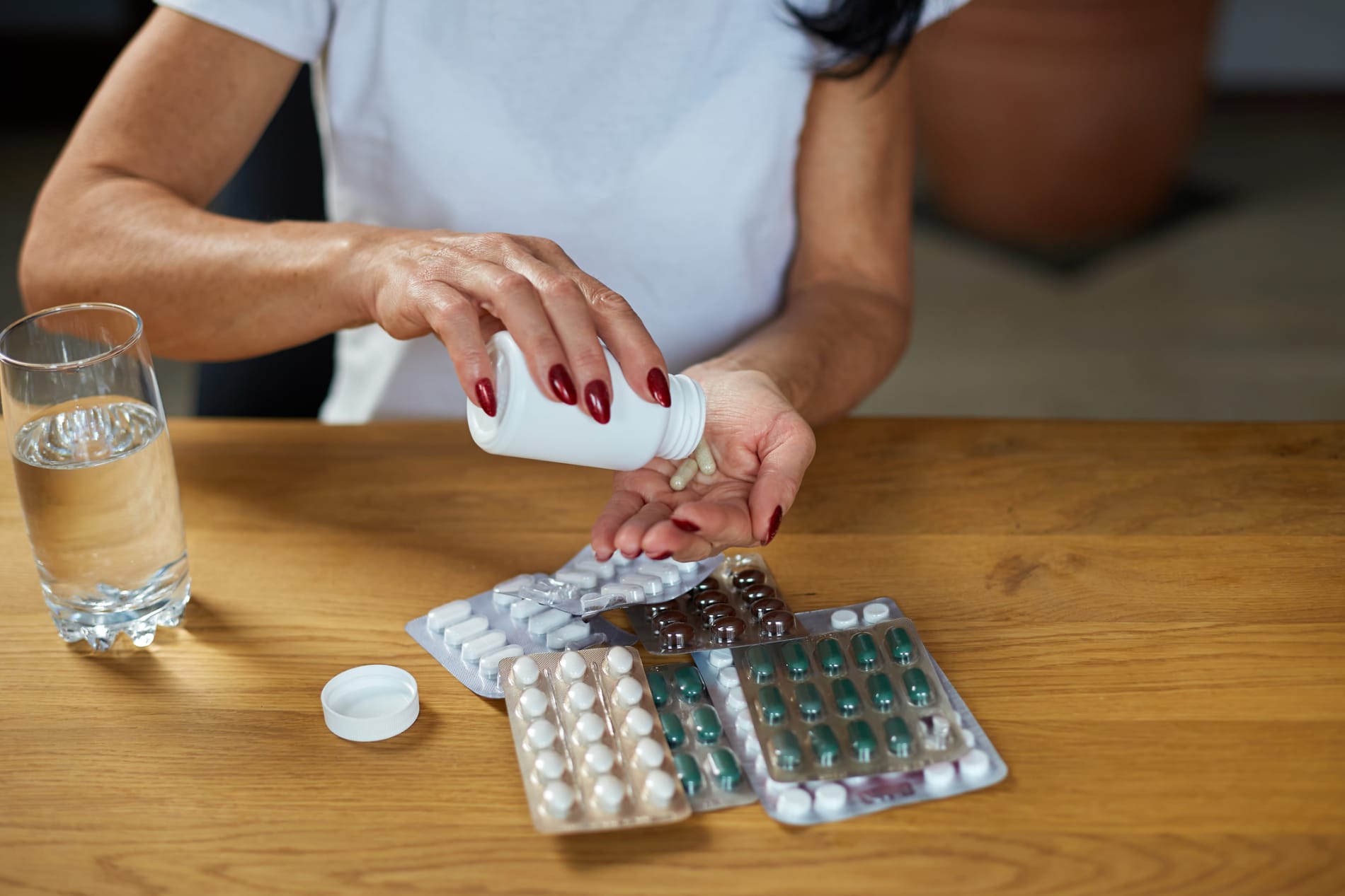 A mature woman sitting at a table with a glass of water, taking a pill from the assorted packs in front of her. 