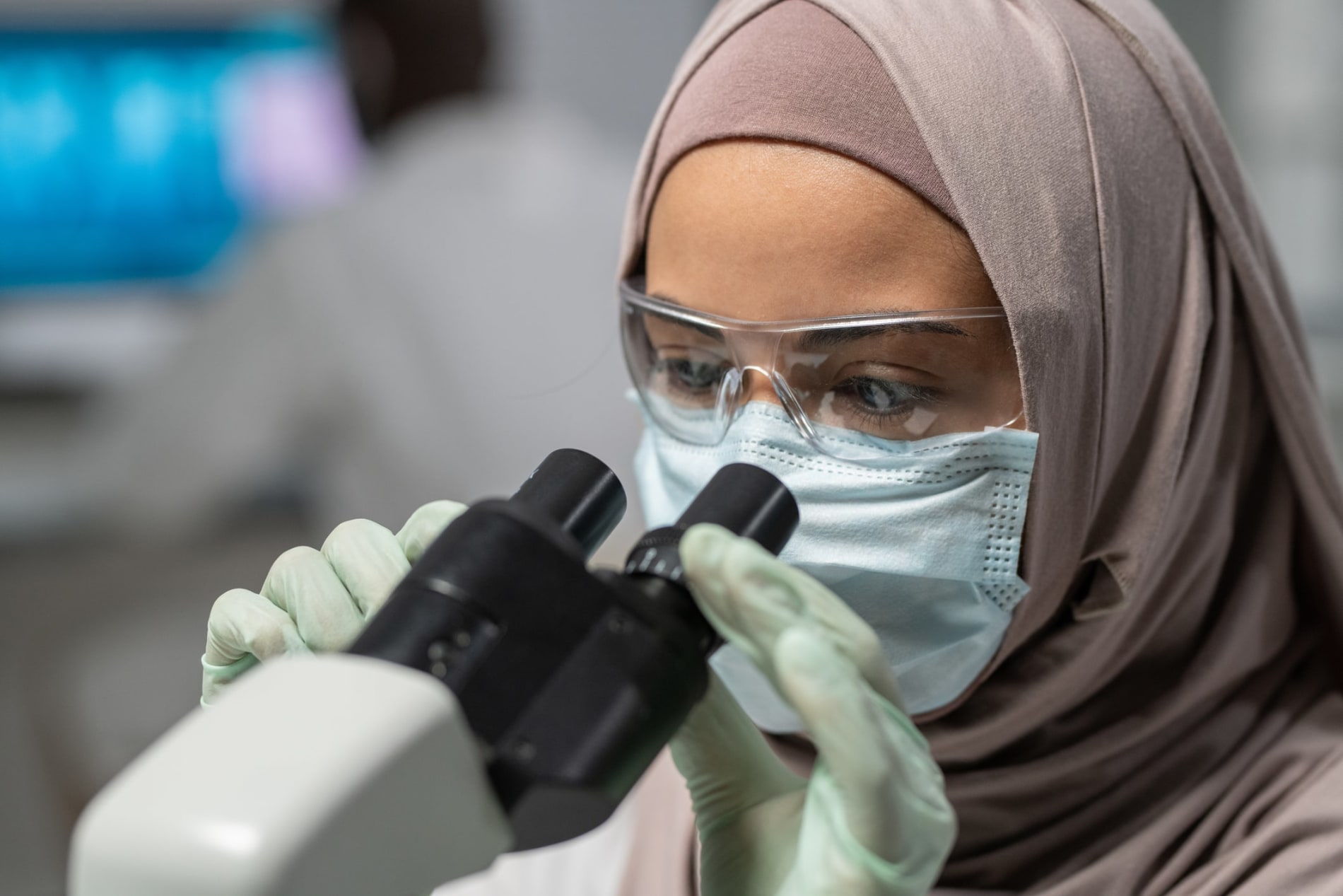 Muslim woman in a hijab, wearing medical gloves and mask, looking into a microscope