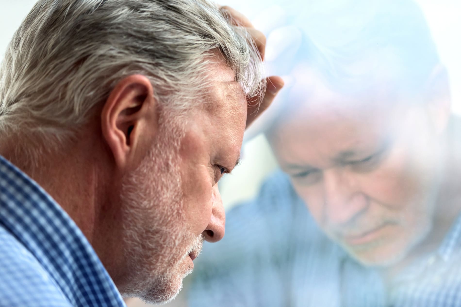 Older man stares out a window with his hand on his forehead staring out a window.