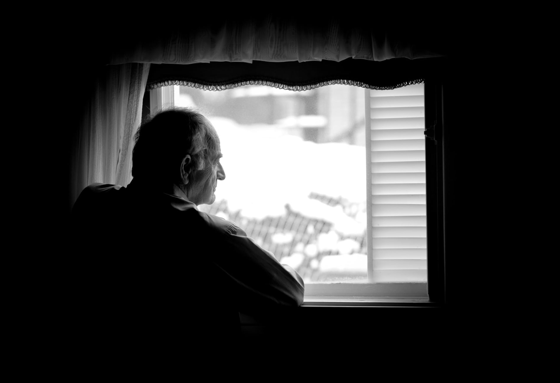 Black-and-white shot of a lonely elderly man looking out a window