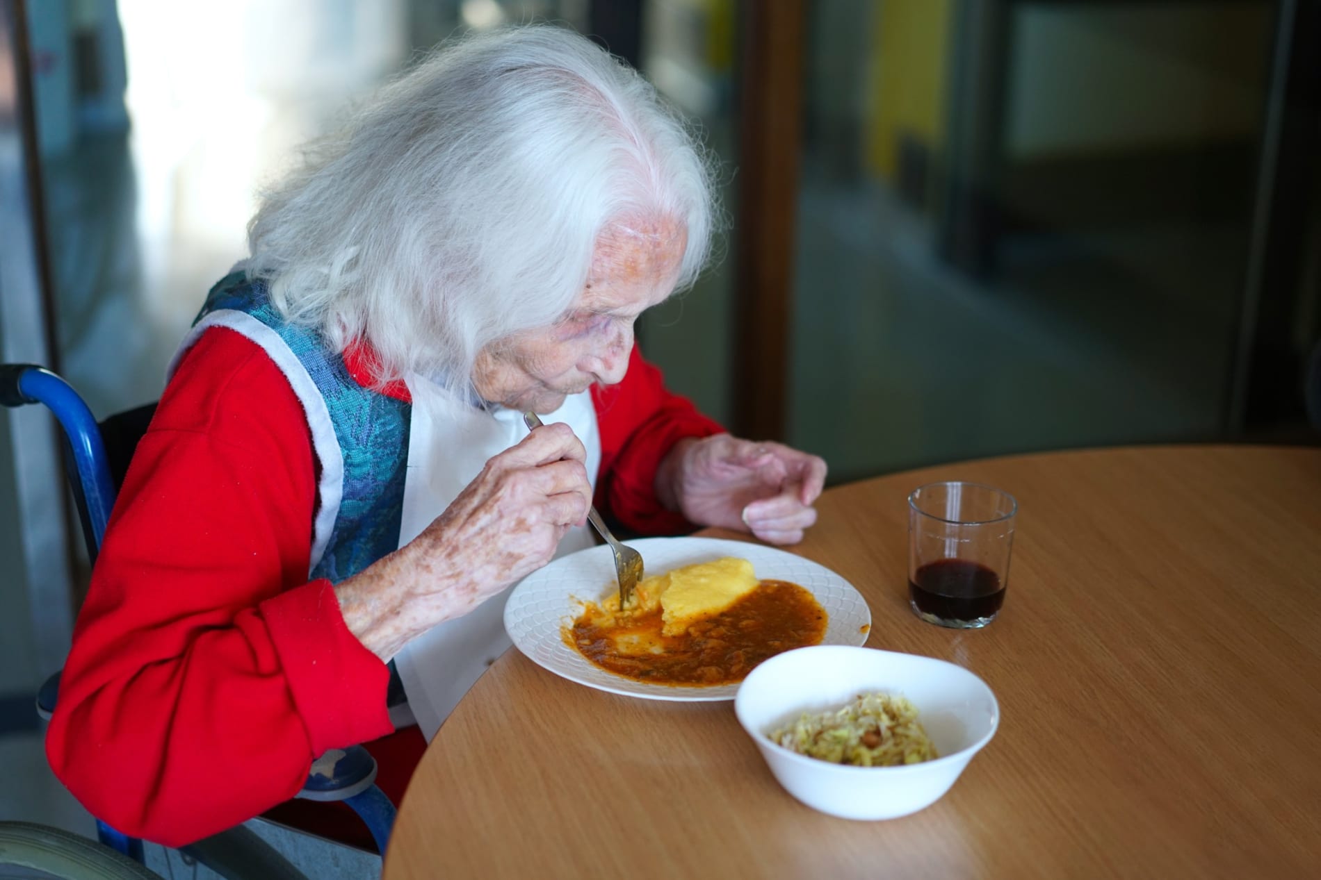 An old woman eats a meal at a table, with a bib on.