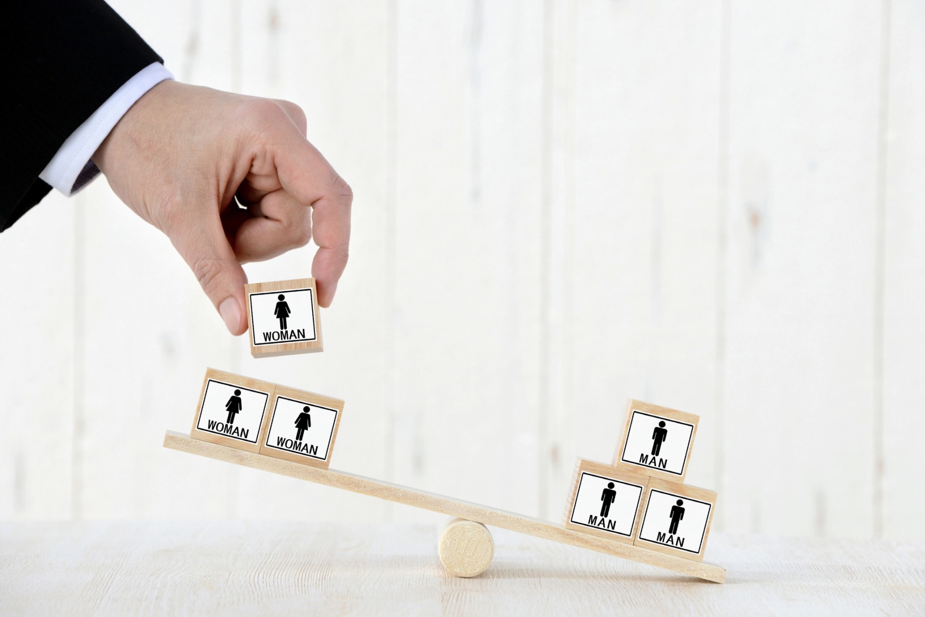 Businessman placing a wooden block with the outline of a woman on a seesaw, thinking about balance between men and women. 