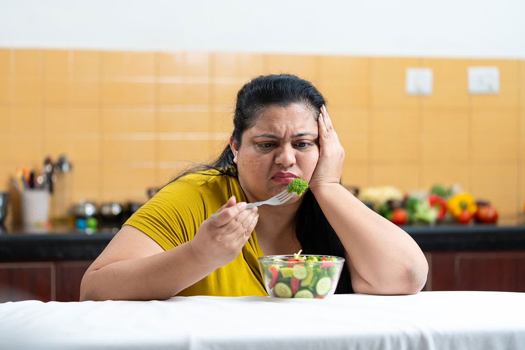 A middle aged woman with her elbows on a dining table looks sadly at food in front of her.