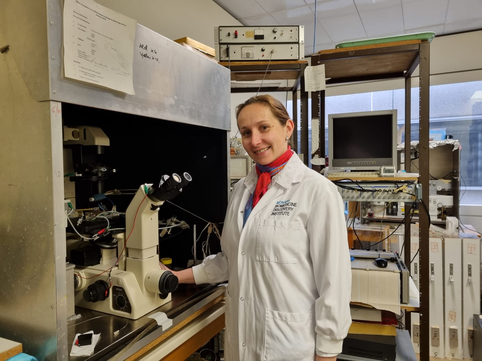 A smiling Dr Olesia Moroz stands beside a microscope in a Monash University laborartory.