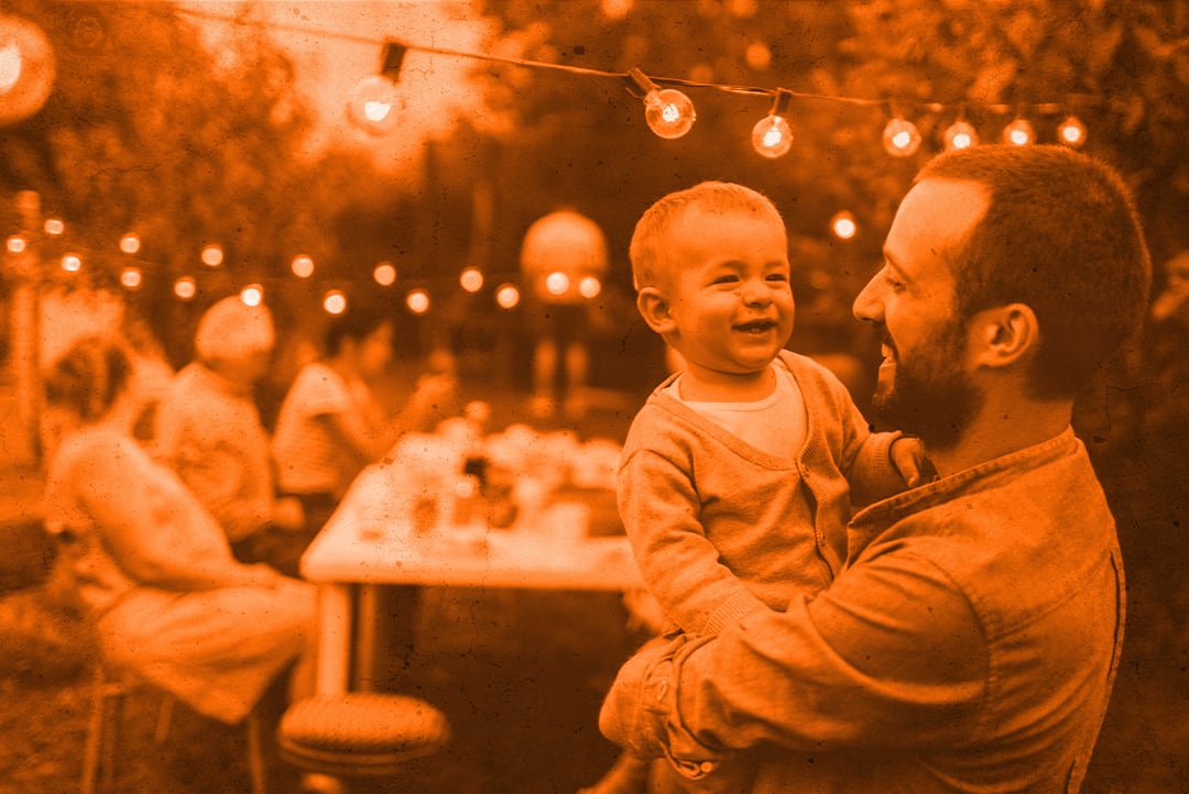 A father with a beard holds his infant son at an outdoor party. There are other partygoers seated at a table in the background, and a string of electric lights over the man's head. The father and baby are both smiling.