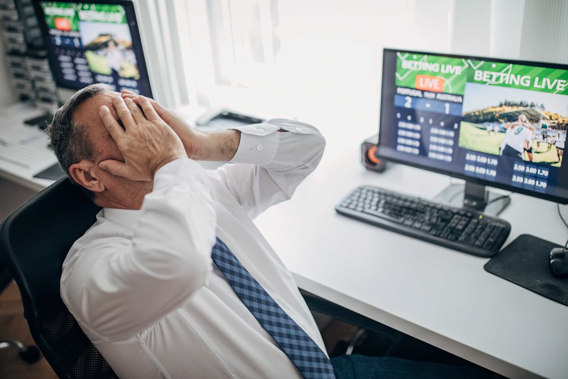 distressed man at his desk with his head in his hands and a computer with a sport betting account.