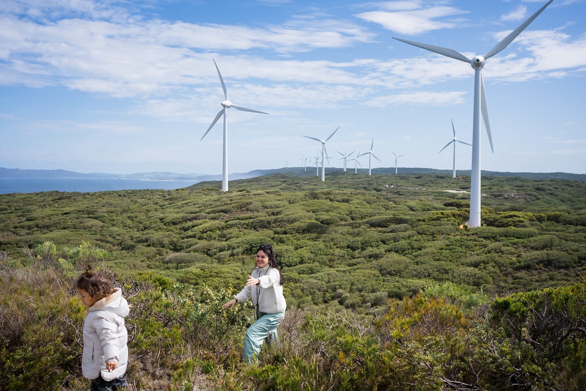 A woman and young girl in coastal bushland in the front of several wind turbines in the distance.