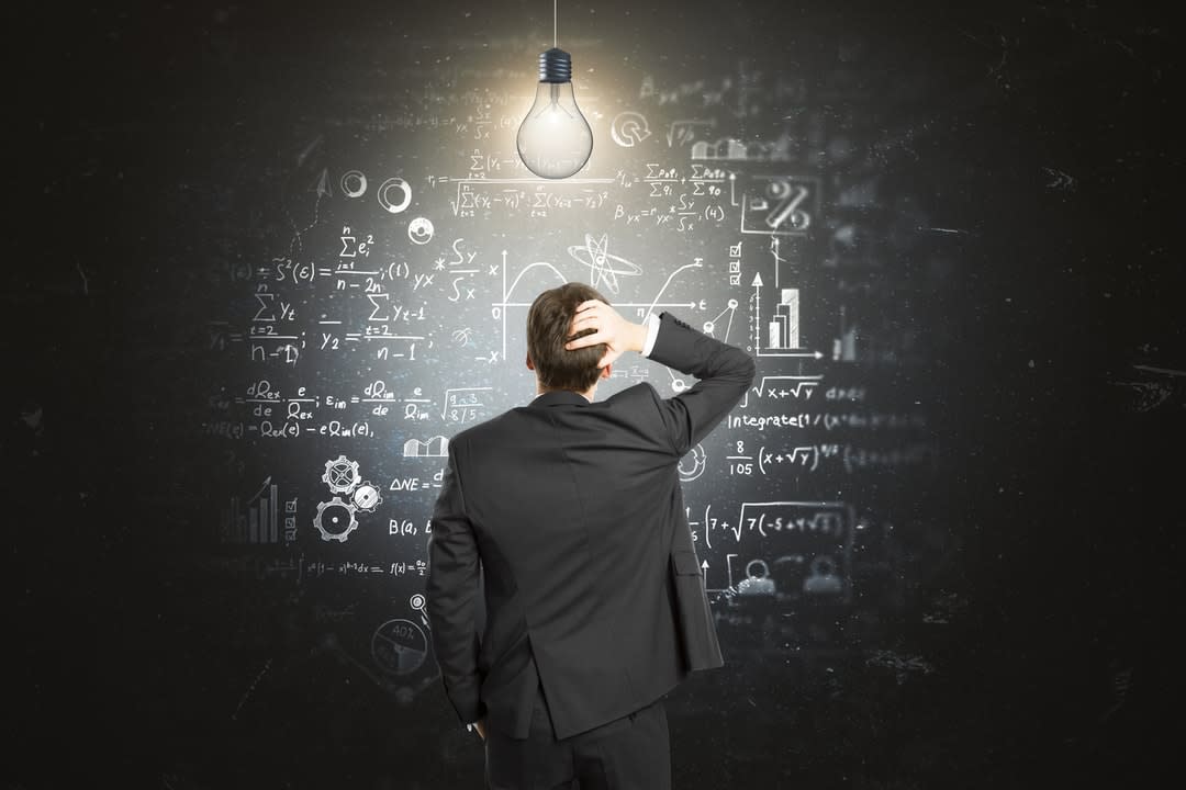 A man stands in front of a blackboard with formulas written in scratching his head and a light bulb illuminating him.