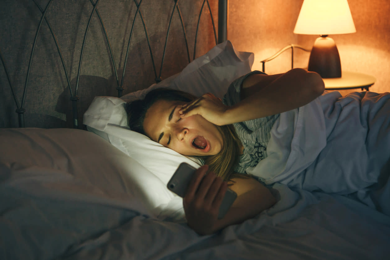 Woman in bed yawning while looking at her phone