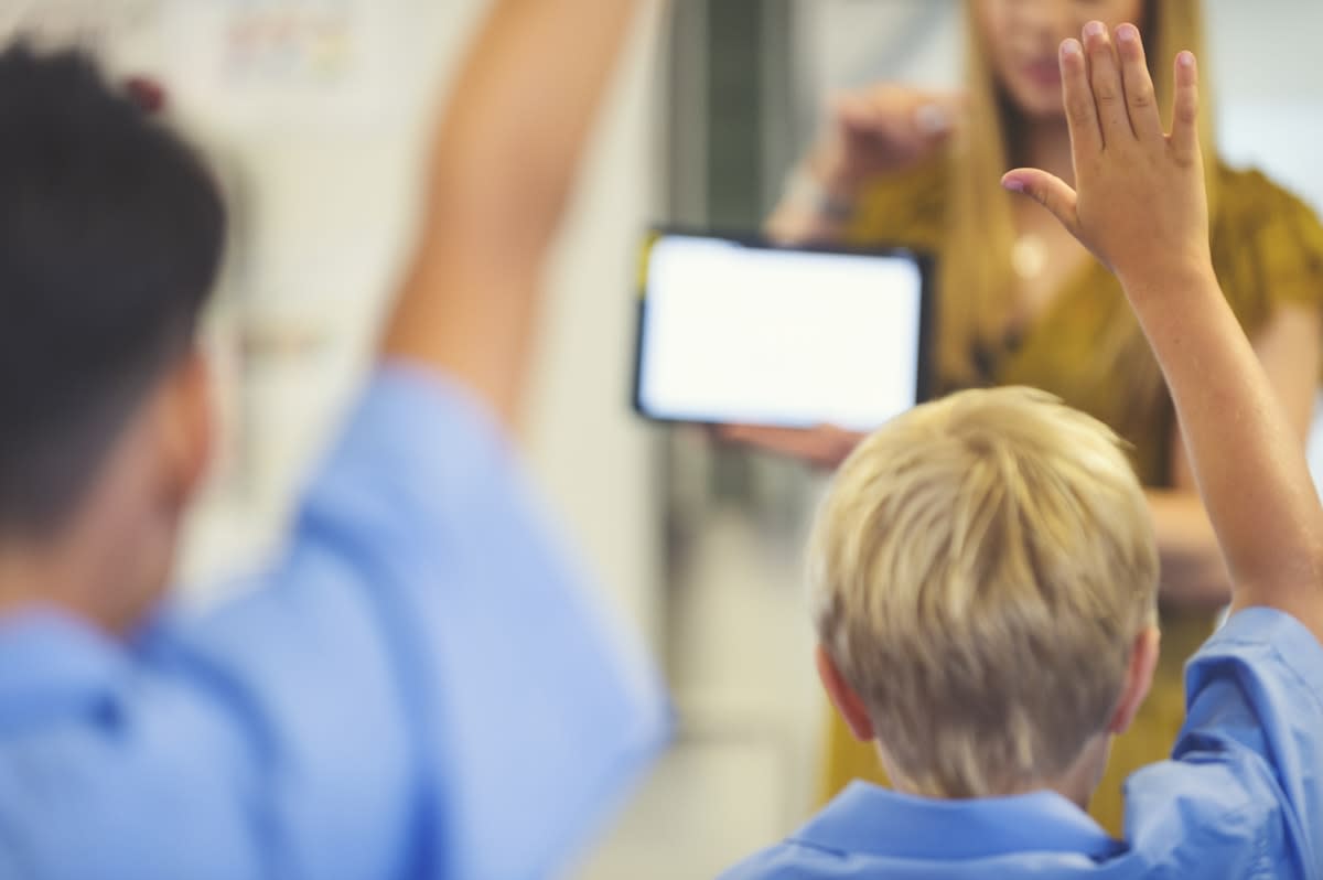 Kids with hands up in a classroom, with the teacher in the background holding up an iPad/tablet