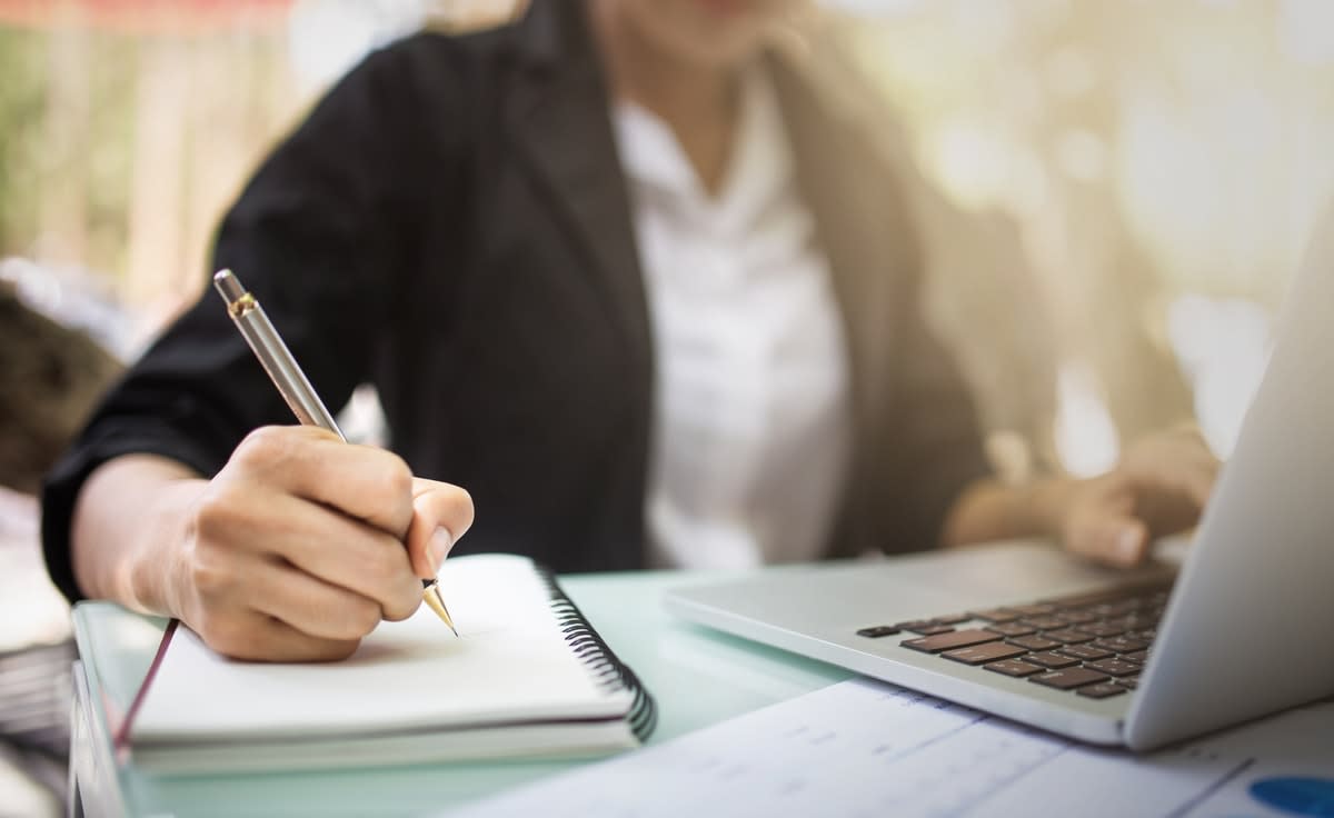 Close-up of a woman writing in a pad, with a laptop in front of her