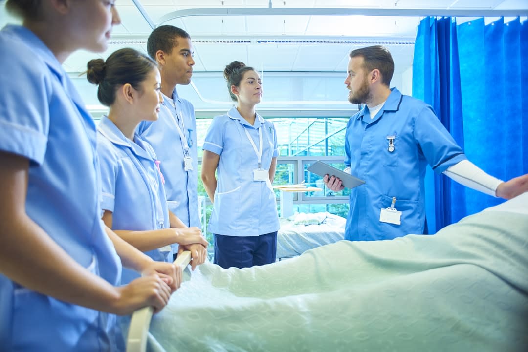 Medical students stand around a bed being given instruction.